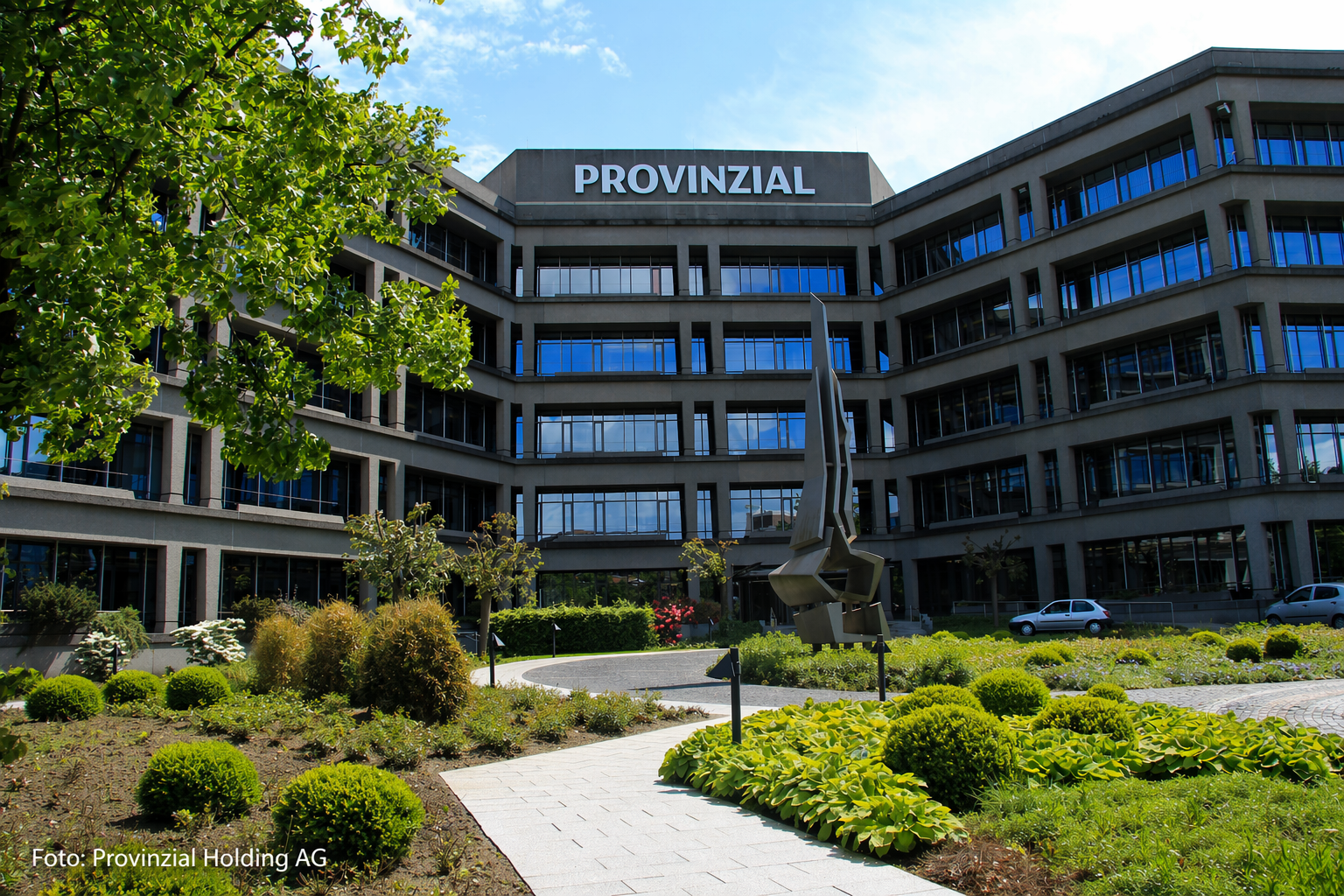 Modern office building with multiple glass windows, a metal sculpture in a landscaped courtyard, under a blue sunny sky.