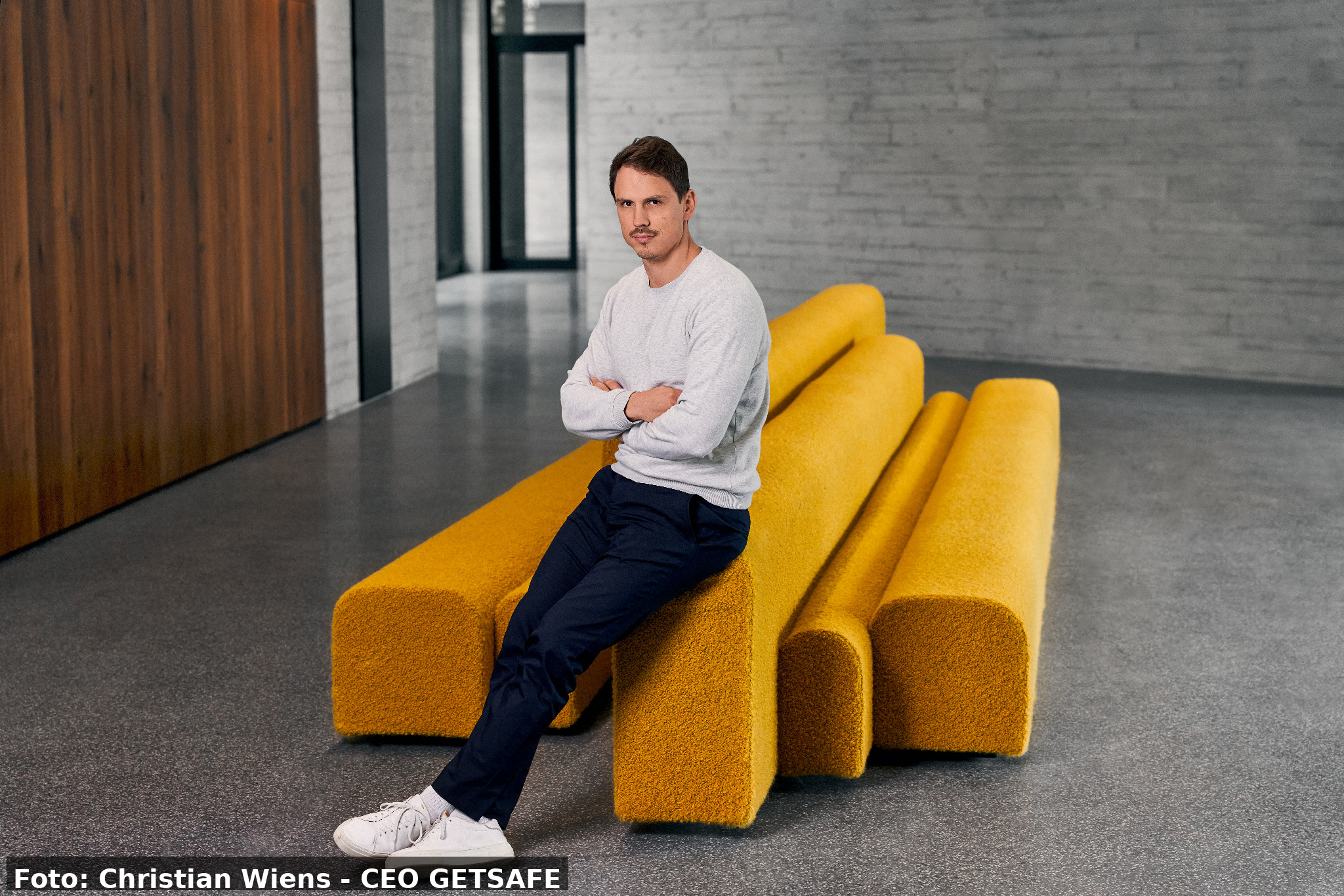 Man seated with arms crossed on bright yellow modular benches in a modern, minimalist hallway.